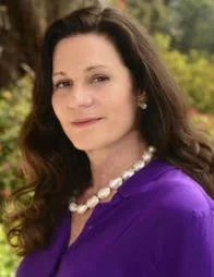 A woman with long dark hair wearing a purple top and pearl necklace outdoors with trees in the background.