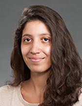 Portrait of a young woman with wavy brown hair, wearing a beige top, smiling softly against a neutral gray background.