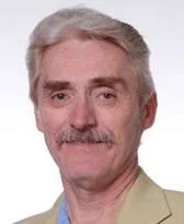 A portrait of a middle-aged man with gray hair, a mustache, and light skin, wearing a beige blazer over a blue shirt.
