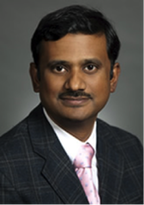 Professional headshot of a man with dark hair, wearing a navy checkered suit and pink tie, against a dark background.
