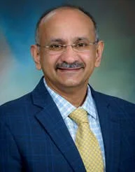 A professional headshot of a smiling man wearing glasses, a blue suit, a light blue checkered shirt, and a yellow tie, against a green and blue background.