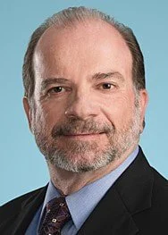A middle-aged man with a beard and receding hairline, dressed in a dark suit, light blue shirt, and patterned tie, posing against a light blue background.