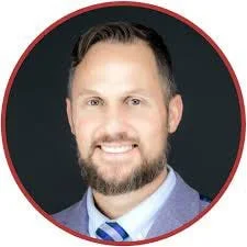 Headshot of a smiling man with a beard, short dark hair, wearing a suit and tie, against a dark background