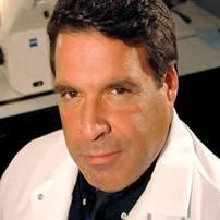 A man with dark hair wearing a white coat, possibly a doctor or scientist, looking at the camera.