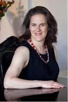 A woman smiling with dark, curly hair, wearing a black sleeveless top and a necklace, sitting at a desk or table.