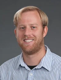 A young man with blond hair, a beard, and a mustache wearing a light blue striped shirt and smiling against a gray background.