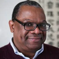 A middle-aged man with glasses and short hair, wearing a dark sweater over a collared shirt, smiling in front of a blurred background.