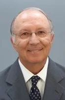A man with glasses, wearing a dark suit, white shirt, and a patterned tie, smiling at the camera.