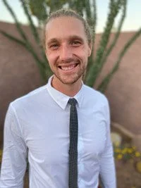 Portrait of a smiling man in a white shirt and tie, standing outdoors with a tree and a brown wall in the background.