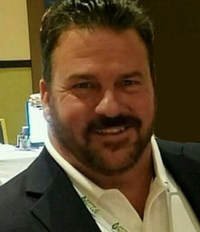 Man with dark hair and beard smiling, wearing a dark blazer and white shirt, indoor setting.