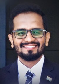 Portrait of a smiling man with glasses, a beard, wearing a suit and tie, against a dark background.