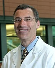 A male doctor smiling, wearing glasses, a white coat, and a patterned tie, standing in a modern clinical or hospital environment.