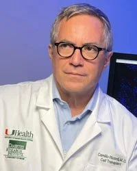 A man wearing glasses and a white medical coat, standing in front of a blue background with a computer monitor.