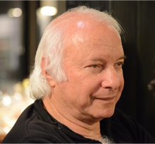 A close-up side profile of an older man with white hair and light skin, wearing a dark shirt.