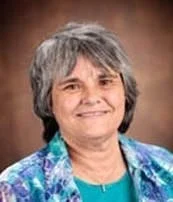 A smiling woman with short gray hair, wearing a blue patterned shirt and a teal top, standing against a brown background.
