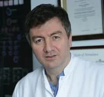 A man in a lab coat standing in a medical or laboratory setting.