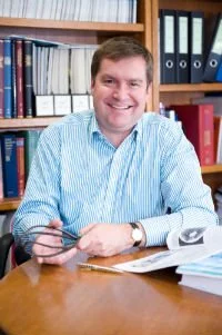 A man smiling, sitting at a wooden desk with papers and a pen, in front of a bookshelf filled with binders and books.