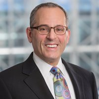 A man wearing glasses, a dark suit, a light-colored shirt, and a colorful tie, smiling in an office setting with large windows.