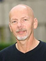 A middle-aged man with a bald head, gray beard, and mustache, wearing a black shirt, standing outdoors with blurred greenery in the background.