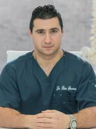 Young man in navy medical scrubs sitting at a desk, looking at the camera.
