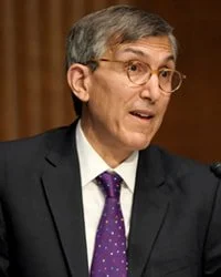 A man with gray hair and glasses wearing a dark suit, white shirt, and purple tie, speaking or gesturing in a formal setting.