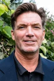 A man with dark hair and a beard, wearing a dark blazer over a black shirt, standing outdoors with greenery in the background.