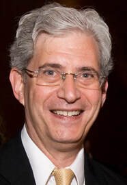 A smiling middle-aged man with gray hair, glasses, wearing a black suit, white shirt, and a yellow tie.