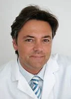 Portrait of a male doctor in a white coat and blue striped tie, smiling at the camera.