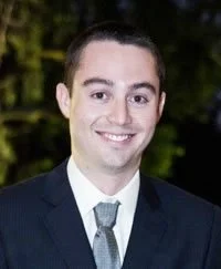 A young man smiling, wearing a dark suit and tie, outdoors with trees in the background.