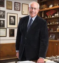 A smiling older man in a dark suit standing in a room with framed photographs and awards on the wall behind him.