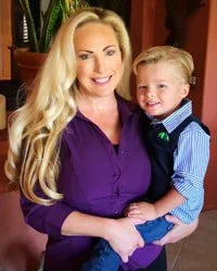 A woman with long blonde hair smiling and holding a young boy with blonde hair, both dressed in casual clothing indoors.