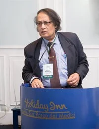 A man with glasses and shoulder-length hair, wearing a dark suit and tie, standing behind a blue podium labeled 'Holiday Inn'.