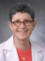 A woman with short curly hair, wearing glasses, a white lab coat, and a red shirt, smiling.