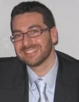 A man with glasses, dark hair, and a beard, smiling, dressed in a suit and tie.