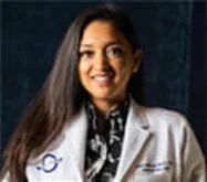A woman with long dark hair in a white medical coat, smiling, in a professional portrait.