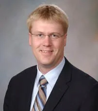 A young man with blonde hair, glasses, wearing a dark suit, light blue shirt, and striped tie, smiling against a plain background.