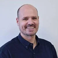 A smiling man with a beard and a shaved head, wearing a dark shirt, standing against a plain light-colored background.