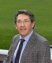 A man in a gray checked suit with glasses standing outdoors on a grassy area.