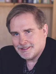 Close-up of a middle-aged man with light brown hair and a beard, smiling in an indoor setting.