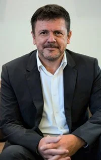 A man with short dark hair and a beard, wearing a dark blazer and white shirt, sitting against a plain light-colored background.