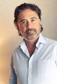 A middle-aged man with dark hair and a beard, wearing a light blue button-up shirt, standing against a neutral background.