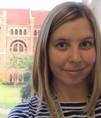 A woman with shoulder-length blonde hair smiling, standing outdoors with a historic building and palm trees in the background.