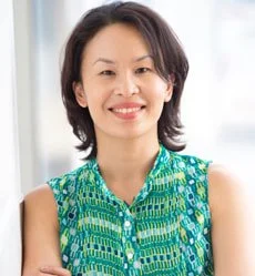 Smiling woman with shoulder-length dark hair wearing a green and blue patterned sleeveless top, standing indoors.