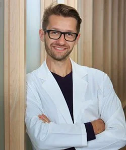 A smiling man wearing glasses and a white lab coat, standing with arms crossed against a wooden background.