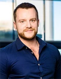 Portrait of a man with short hair and a beard, wearing a navy blue button-up shirt, smiling in front of a window.