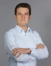A young man with dark hair wearing a light blue button-up shirt with arms crossed, standing against a neutral gray background.