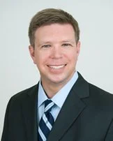 Portrait of a young man in a suit smiling against a light background.