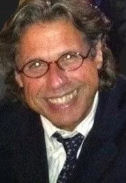 A smiling man with curly dark hair, glasses, and a patterned tie, wearing a dark suit jacket.