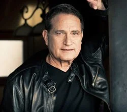 A middle-aged man with short dark hair, wearing a black leather jacket, posing indoors with one arm raised.