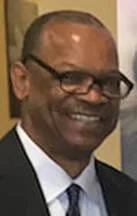 A smiling man wearing glasses, a dark suit, white shirt, and blue tie.